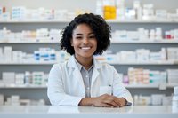 African American female pharmacist with curly hair and bright smile standing at pharmacy counter with medicine shelves in background, wearing white lab coat.