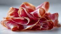 Thinly sliced prosciutto ham arranged in elegant folds on white surface, showcasing rich pink and red colors with marbled fat streaks for culinary presentations.