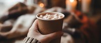 Hand holding a kraft paper cup of hot cocoa with marshmallows, cozy warm background with soft bokeh lights, winter comfort drink.