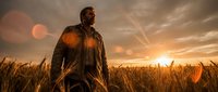 Man with beard wearing brown jacket standing in golden wheat field at sunset with warm sky and sun flare, suitable for agriculture, lifestyle and inspirational content.
