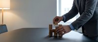 Professional businessman in dark suit arranging wooden building blocks on modern office desk, strategic planning and problem-solving concept for corporate success.