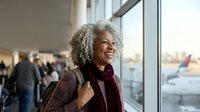 Happy mature African American woman with curly gray hair laughing joyfully at airport terminal window, carrying luggage for travel adventures.