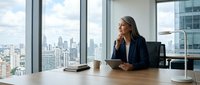 Professional businesswoman in navy blazer sitting at modern office desk with city skyline view through floor-to-ceiling windows during daytime meeting.