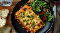 Traditional lasagna with melted cheese and herbs served on black plate alongside fresh garden salad and crusty bread slices, photographed from above.