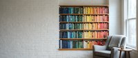 Modern home library interior with colorful rainbow organized bookshelf against white brick wall, comfortable reading chair and natural window light.