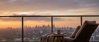 Luxury penthouse interior with comfortable armchair facing floor-to-ceiling windows overlooking sprawling cityscape at golden hour sunset, ideal for real estate and lifestyle marketing.