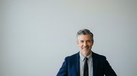 Middle-aged man with gray hair and facial hair wearing navy blazer and gray shirt against white background, suitable for corporate, business, and professional portraits.