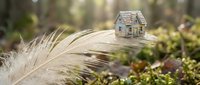 Miniature wooden house model placed on natural feather surrounded by green moss in soft forest light creating dreamy fairy tale atmosphere for home concepts.