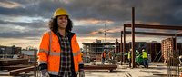 Female construction worker in safety gear smiling at urban building site during sunset with steel framework and crane in background.