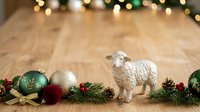 White ceramic sheep figurine surrounded by festive ornaments, evergreen branches, and holiday decorations on wooden surface for seasonal and Christmas design projects.