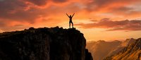 Silhouette of victorious person with raised arms standing on mountain cliff edge against dramatic orange sunset sky over mountain ranges for success concepts.