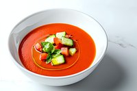 Fresh gazpacho soup garnished with cucumber, tomato and basil in white ceramic bowl on marble background, bright natural lighting.