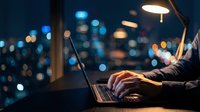 Professional working late at night on laptop with city lights bokeh background creating productive workspace atmosphere for remote work and business.