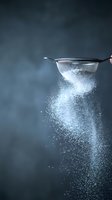 Fine mesh strainer sifting white flour powder creating dramatic dust cloud against dark moody background for culinary and baking concepts.