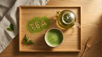 Matcha green tea flat lay on wooden tray with glass teapot, ceramic bowl, and TEA lettering made from powder, top view.