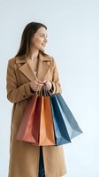 Young woman with long dark hair and fair complexion wearing a tan trench coat, holding multiple colorful shopping bags in red, orange, and blue tones against a white background for retail and consumer lifestyle use.