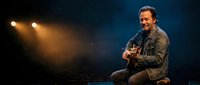 Male musician with acoustic guitar smiling on stage under dramatic spotlights, performing solo concert in dark atmospheric venue.