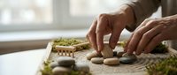 Hands arranging smooth stones in zen garden with rake patterns in sand for meditation and mindfulness practice at home.