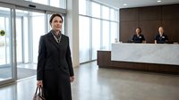 Professional Asian businesswoman in elegant black coat standing in modern hotel lobby with marble reception desk and staff in background.