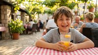 Happy young boy enjoying fresh orange juice at outdoor restaurant terrace during summer vacation with family in Mediterranean setting.