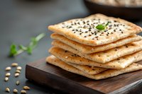 Stack of crispy crackers with sesame and poppy seeds garnished with fresh herbs on wooden cutting board, close up view with selective focus.