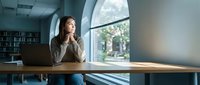 Young professional woman sitting at wooden desk in modern office space with large windows, natural lighting and bookshelves in background for business concepts.