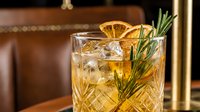 Whiskey cocktail with dried orange slice and rosemary garnish in crystal glass on wooden bar counter in upscale restaurant setting.