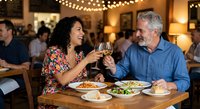 Mature couple enjoying romantic dinner date at upscale restaurant, toasting with wine glasses while sharing meal together in warm ambient lighting.