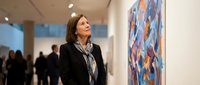 Middle-aged Caucasian woman in black blazer admiring colorful abstract artwork in modern art gallery with visitors in background.