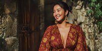 Young Asian woman in burgundy floral dress smiling outdoors against rustic stone wall background with natural lighting for lifestyle and fashion content.