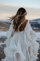 Young woman in flowing white dress with windswept hair stands against mountain landscape at dusk, back turned to camera showing dramatic open-back gown design.