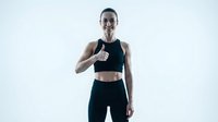 Young athletic woman with short dark hair and glasses wearing black sports bra and leggings, giving thumbs up gesture on white background for fitness and wellness content.