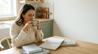 Woman with dark hair and olive complexion, wearing a beige cardigan, sipping from a mug while working at a wooden desk with notebooks and papers, suitable for lifestyle and productivity content.