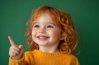 Happy redhead toddler girl giving thumbs up gesture wearing yellow sweater on green background. Cheerful child showing positive emotion and approval sign.