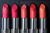 Crushed lipstick bullets in gradient shades from pink to coral arranged in row on dark background, showing texture detail in metallic silver cases for beauty marketing.