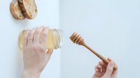 Caucasian woman's hand holding glass jar of honey with wooden dipper and bread slices on white background, food styling for culinary and recipe content.