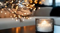 Glowing candle in textured glass holder with blurred fairy lights creating magical bokeh background, cozy winter holiday atmosphere for festive decor.