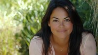 Young Hispanic woman with long dark hair smiling outdoors in natural sunlight against green foliage background, wearing casual attire and delicate necklace.