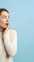 Young woman with brown hair and fair complexion wearing cream sweater, hand to face with surprised expression against blue background.