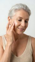 Mature woman applying face cream for anti-aging skincare routine, touching cheek gently with natural smile showing healthy aging and self-care habits.