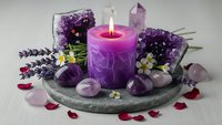 Purple candle burning surrounded by amethyst crystals, lavender flowers and rose petals on stone plate for spa relaxation and meditation rituals.