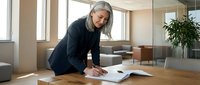 Mature businesswoman with gray hair writing documents at wooden desk in modern office with natural lighting and contemporary furniture.