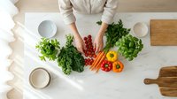 Chef preparing fresh vegetables and salad ingredients on marble countertop with kale, carrots, and bell peppers for healthy cooking and meal preparation.