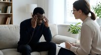 Professional therapist counseling stressed African American businessman during mental health session in modern office setting.
