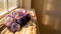 Purple amethyst crystals and black tourmaline gemstones arranged on rustic wooden surface with warm natural sunlight streaming through window.