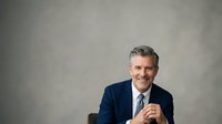 Middle-aged man with gray hair and light skin tone wearing a dark navy blazer and light blue dress shirt with tie, smiling confidently at camera in professional studio portrait setting.