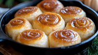 Freshly baked cinnamon rolls with golden brown swirls in cast iron skillet, glazed with sweet sticky caramel sauce, close up view of homemade pastries.