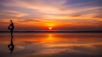 Woman practicing yoga tree pose on beach at sunset with golden sky reflection on wet sand creating peaceful meditation scene for wellness and mindfulness.