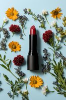 Red lipstick surrounded by yellow daisies, burgundy carnations, and white wildflowers on light blue background. Feminine beauty and natural cosmetics flatlay composition.