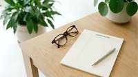 Minimalist wooden desk with notepad, pen, and glasses surrounded by green plants in a bright, clean workspace setting.